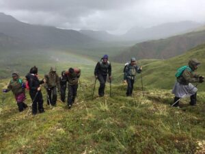 A group of Denali Backcountry Adventures students wear full raingear while hiking in the tundra. They carry trekking poles and while their hoods are up protecting them from the elements, there is sun dotting the ground and coming through the low clouds behind them.